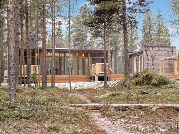 A cabin with a deck and large windows, surrounded by tall trees and a snowy ground.