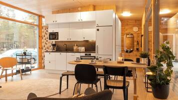 A modern kitchen with white cabinets, a dining area with a wooden table and chairs, and a large window offering a view of a car and trees.