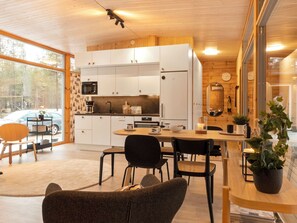 A modern kitchen with white cabinets, a dining area with a wooden table and chairs, and a large window offering a view of a car and trees. - Solokoto by Interhome (Inari)