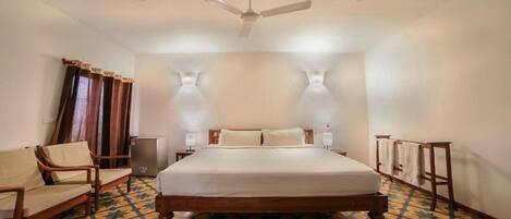 Una habitación de hotel con una cama, un ventilador de techo, una mesita de noche, una lámpara, una silla y un suelo estampado.