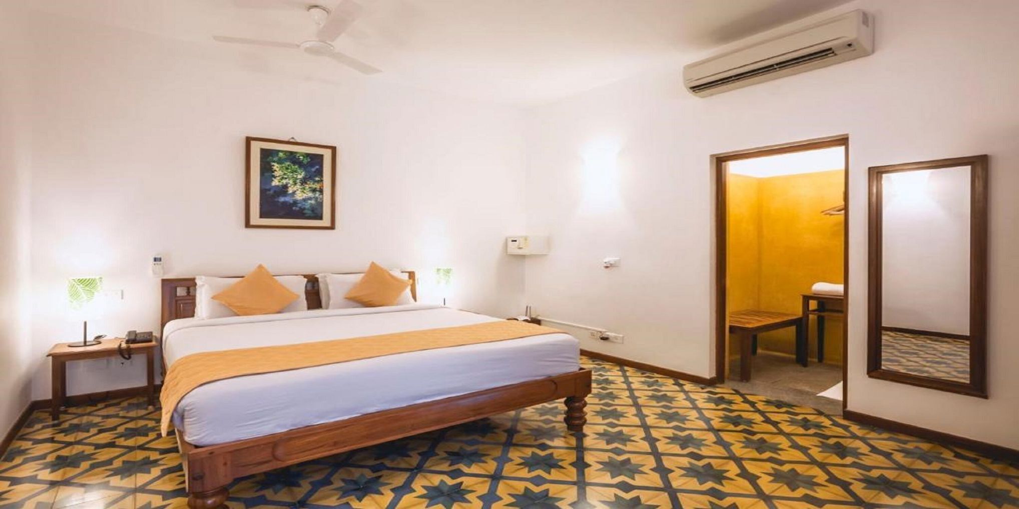 A hotel room with a bed, bedside tables, a ceiling fan, an air conditioning unit, a framed picture on the wall, and a patterned floor.