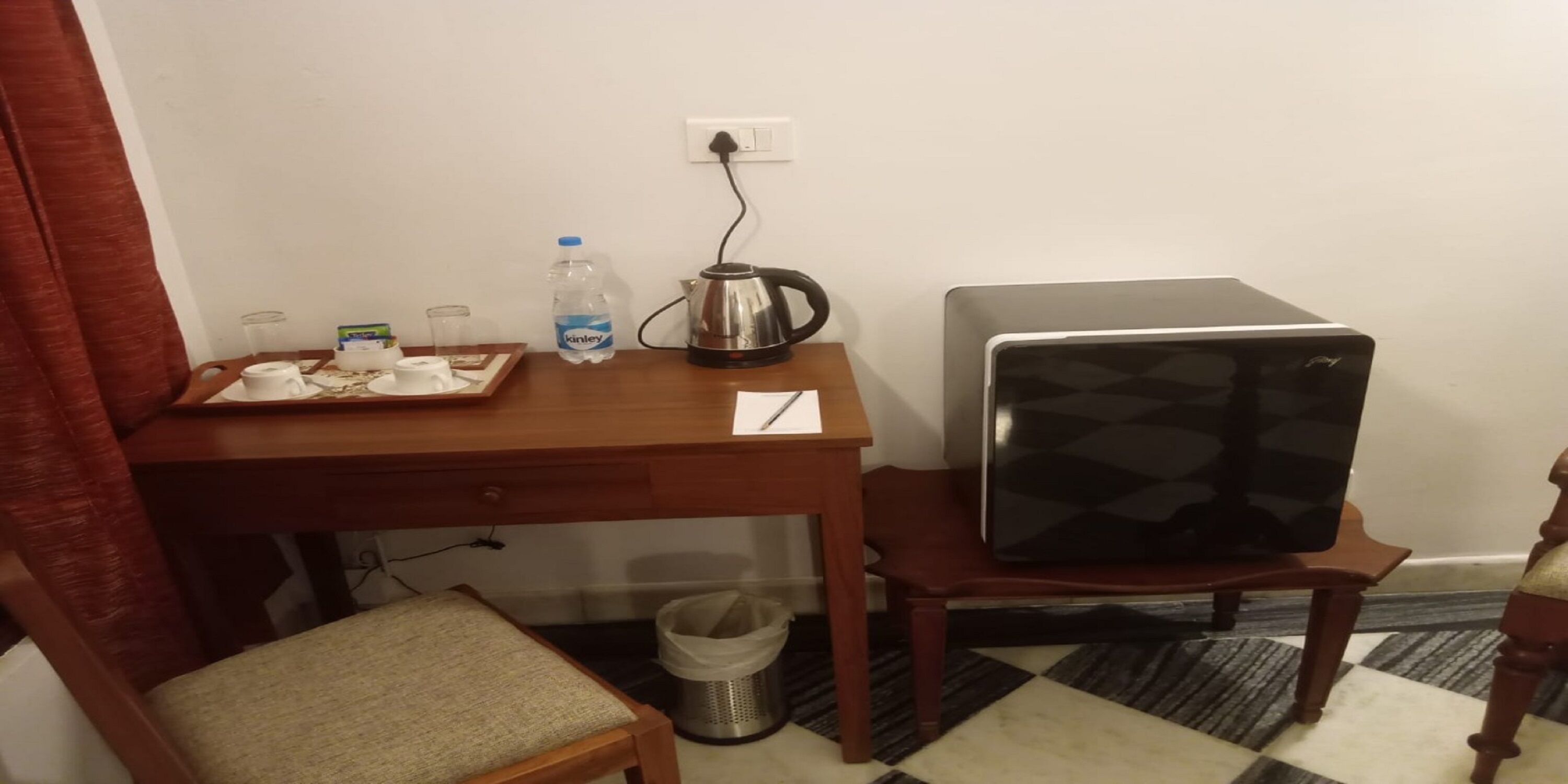 A small room with a wooden desk, a chair, a refrigerator, a kettle, a water bottle, and a trash bin.