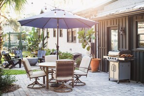 Eine Terrasse im Freien mit einem runden Tisch, vier Stühlen, einem großen Sonnenschirm, einem Grill und einem Blumentopf.