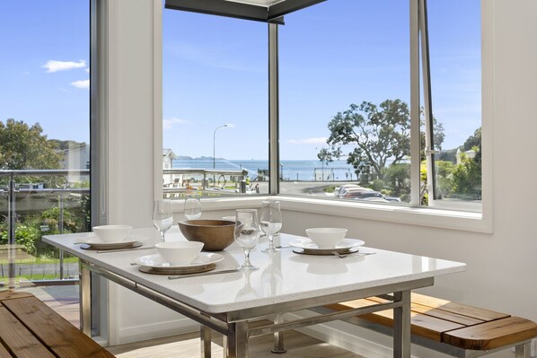 A dining table with white plates, bowls, and wine glasses, set against a large window overlooking a scenic view of the sea and a promenade.
