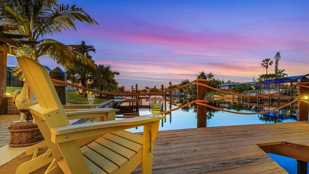 A wooden deck with a lounge chair and a glass of drink, overlooking a calm pool with a rope barrier and palm trees.