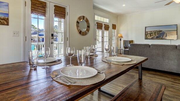Un comedor con una mesa de madera dispuesta para cuatro, un espejo redondo y vistas a una playa a través de las ventanas.