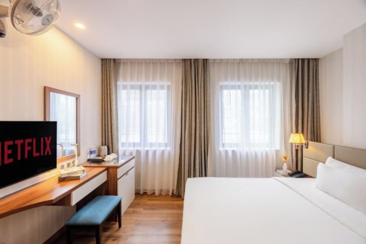 A hotel room with a bed, a desk with a television, a bench, and curtains.