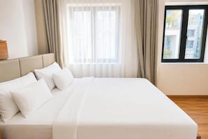 A large bed with white linens and pillows, a wooden headboard, sheer curtains, and a view of buildings through a window.