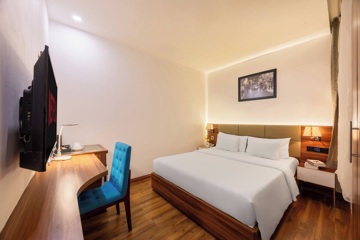 A hotel room with a bed, a desk with a chair, a television, and a bedside table with a lamp.
