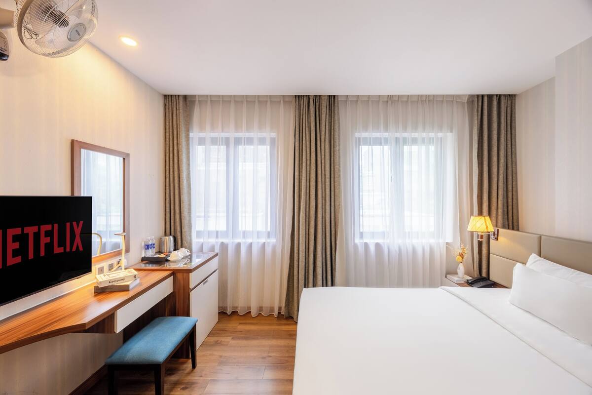 A hotel room with a bed, a desk with a television, a lamp, and a window with curtains.