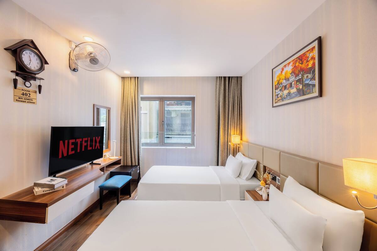 A hotel room with a bed, a desk with a TV displaying 'NETFLIX', a clock, a fan, and a painting on the wall.