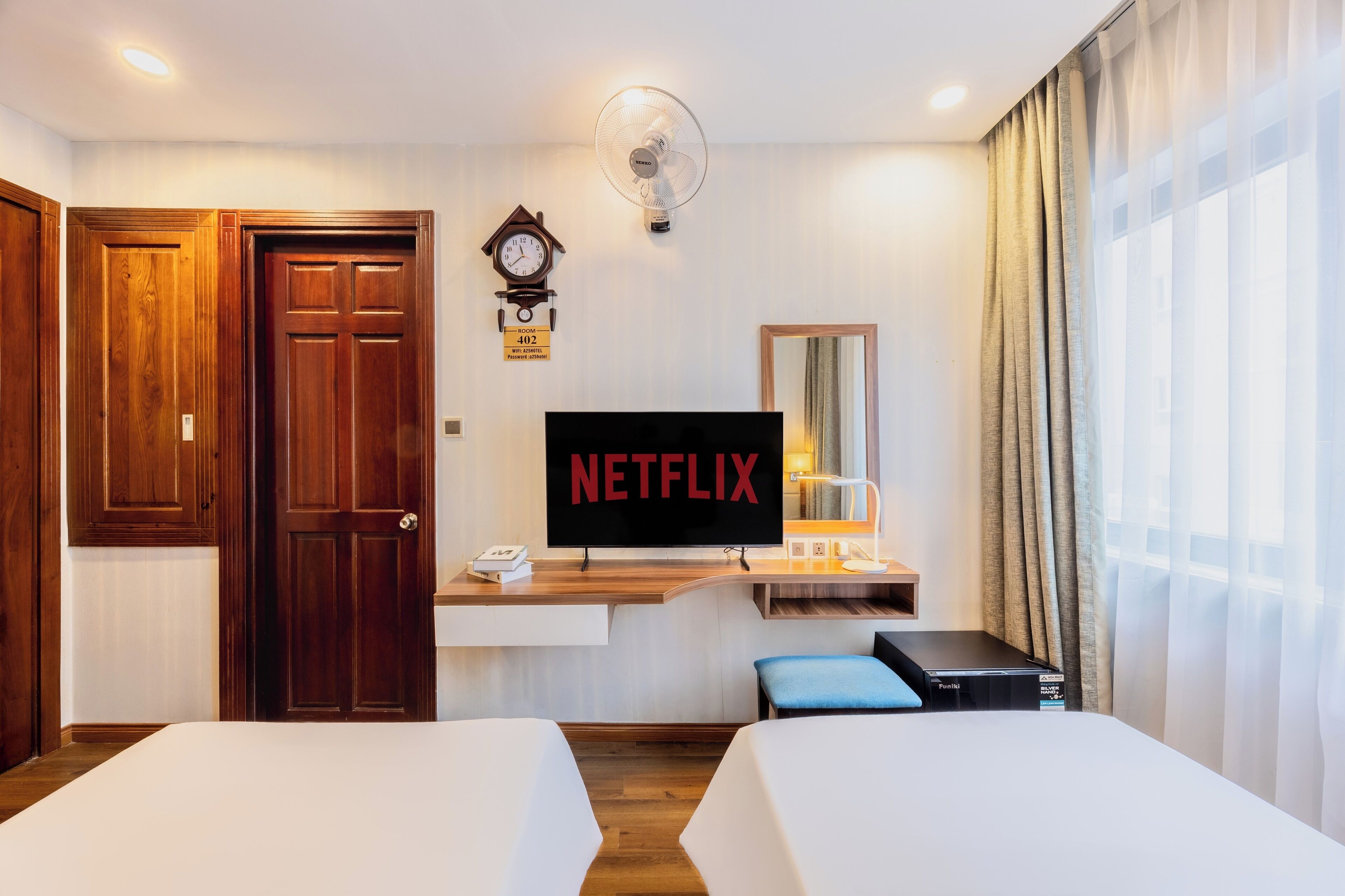 Quarto de hotel com duas camas, uma televisão exibindo o logotipo da Netflix, um relógio e um ventilador.
