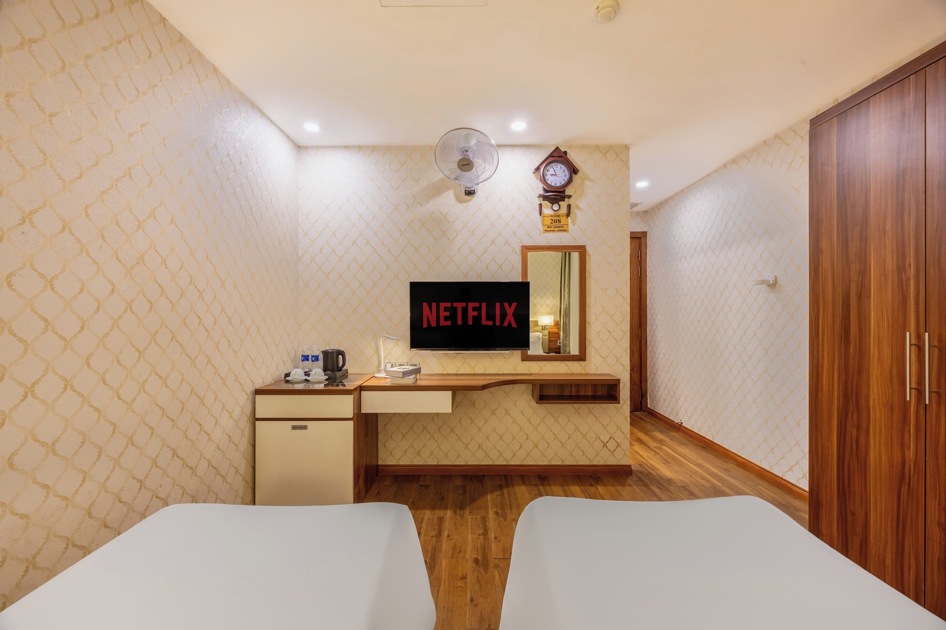 Habitación de hotel con cama, escritorio con televisor mostrando 'NETFLIX', un reloj y un espejo.