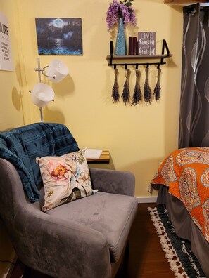 Une pièce chaleureuse avec un fauteuil gris, un coussin décoratif, un tableau représentant un ciel étoilé, une étagère avec un vase et des bougies, et un lit recouvert d’une couverture à motifs.