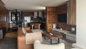 Un salon moderne avec un canapé, des fauteuils et une table basse. Un escalier mène à un espace cuisine équipé d’un réfrigérateur et d’une table à manger.