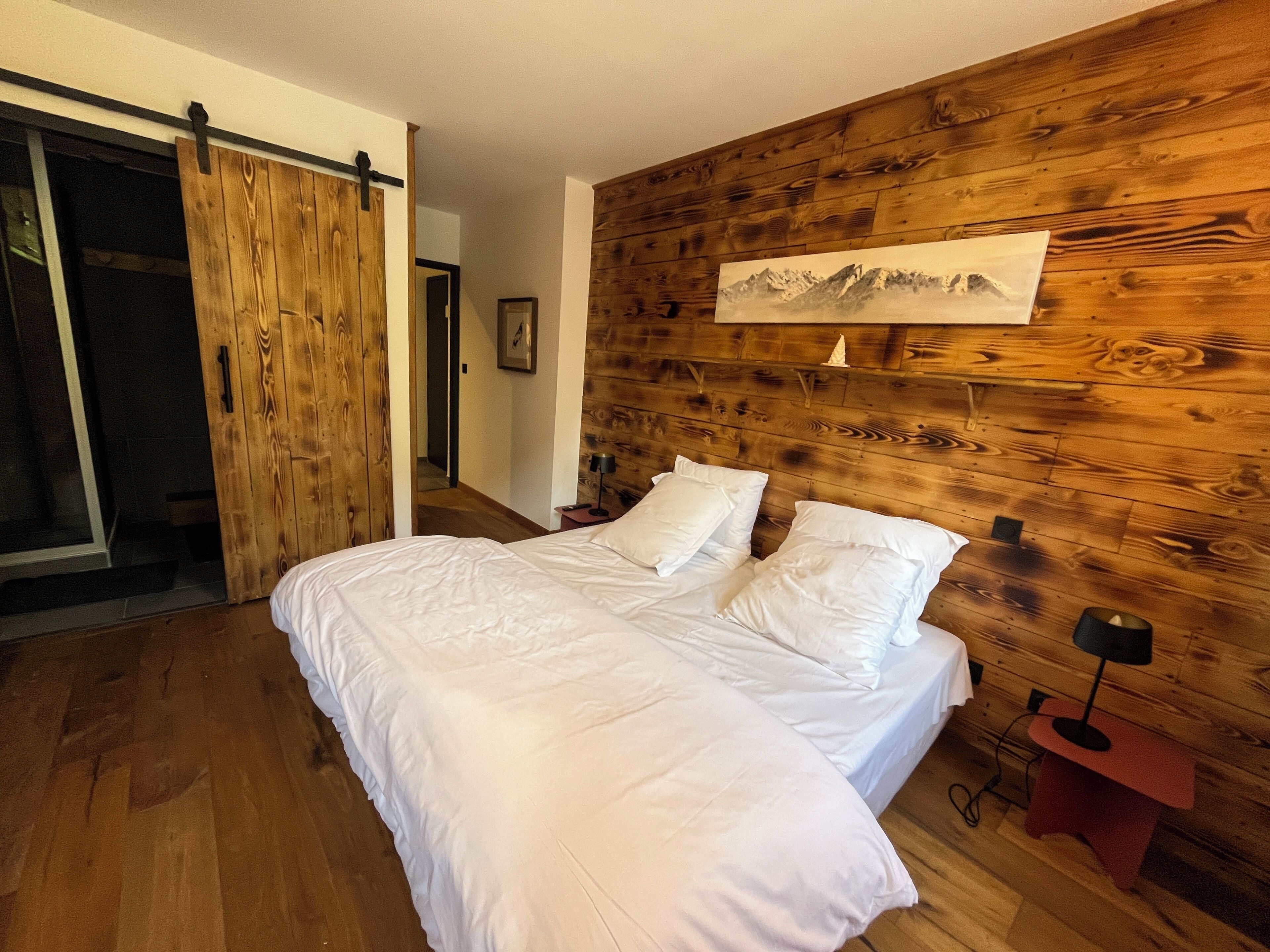A bedroom with a wooden headboard, a bed with white bedding, a nightstand with a lamp, and a sliding door leading to another room.