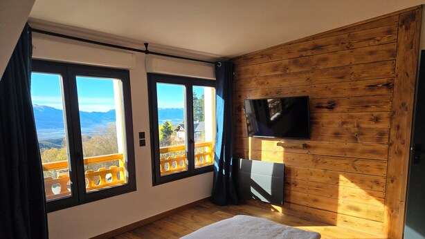 Une chambre dotée d’un lit, d’un téléviseur fixé au mur en bois et de grandes fenêtres donnant sur l’extérieur.