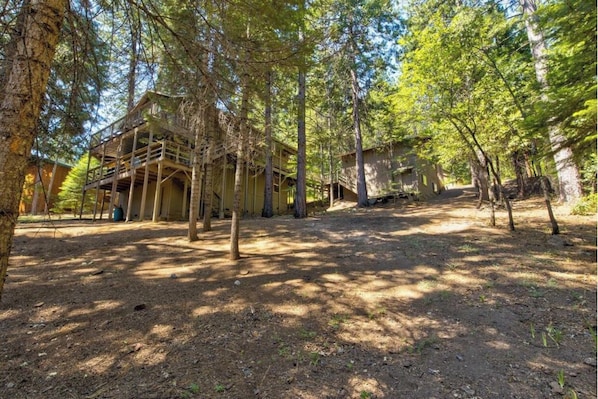 A rustic cabin with a wooden deck and multiple levels, surrounded by tall trees in a forest setting.
