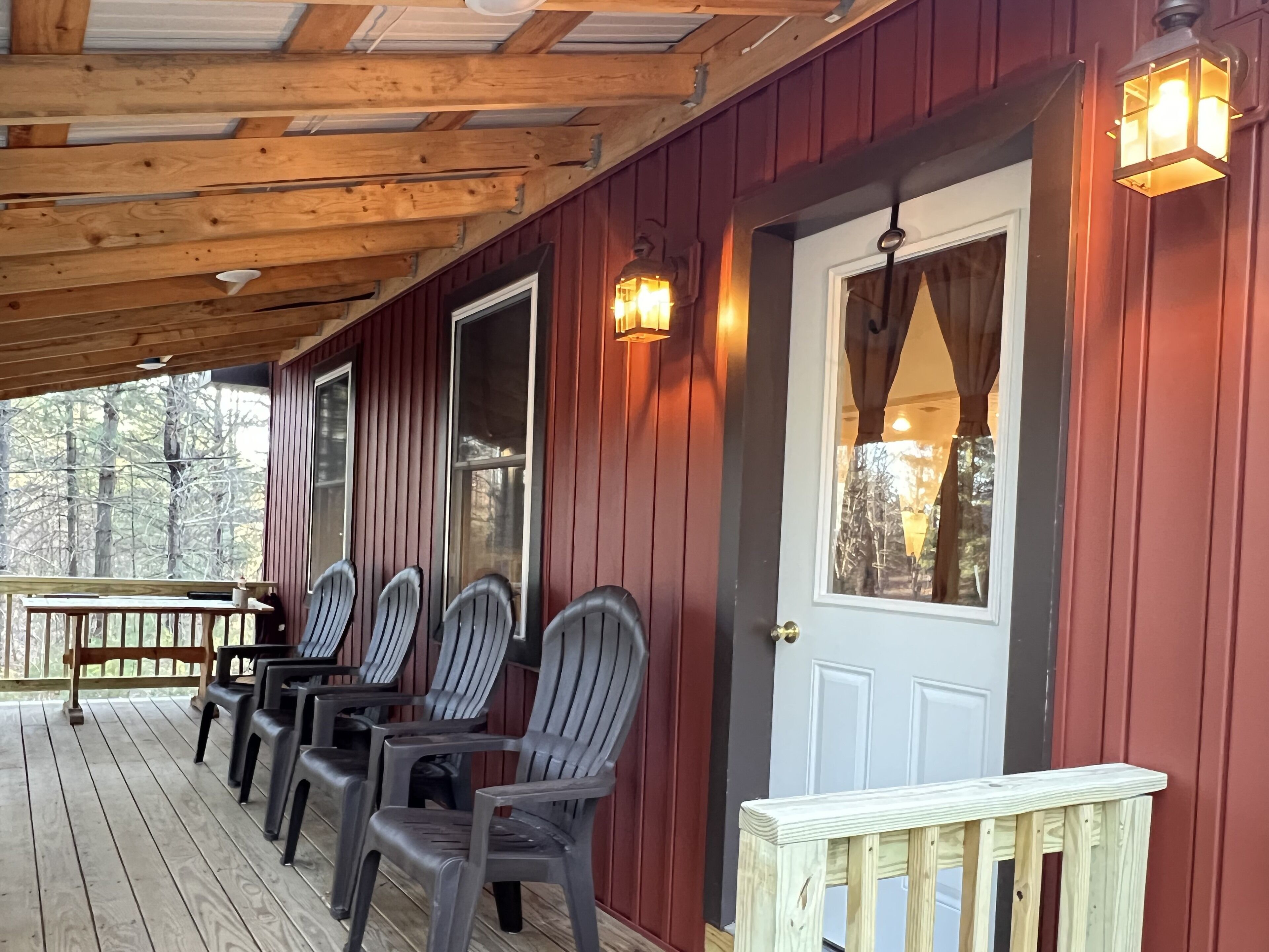 A covered porch with wooden benches and chairs, a white door with a window, and wall-mounted lights.