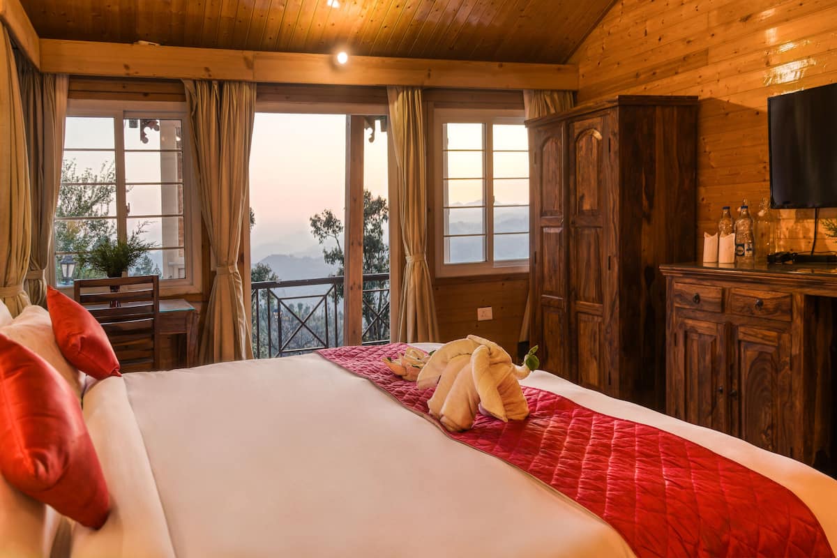 A wooden cabin room with a large bed, a desk, and a balcony with a view of the mountains.