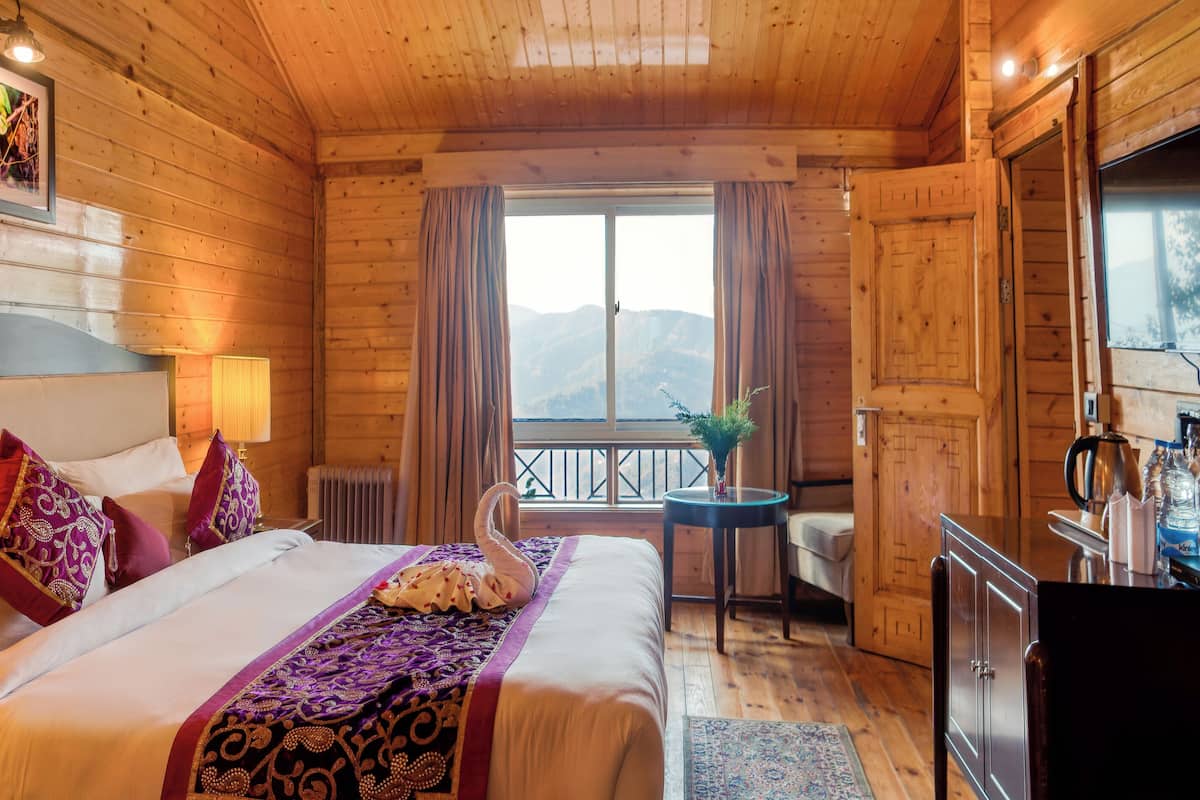 A wooden cabin room with a bed, bedside lamps, a small table, and a view of the mountains.