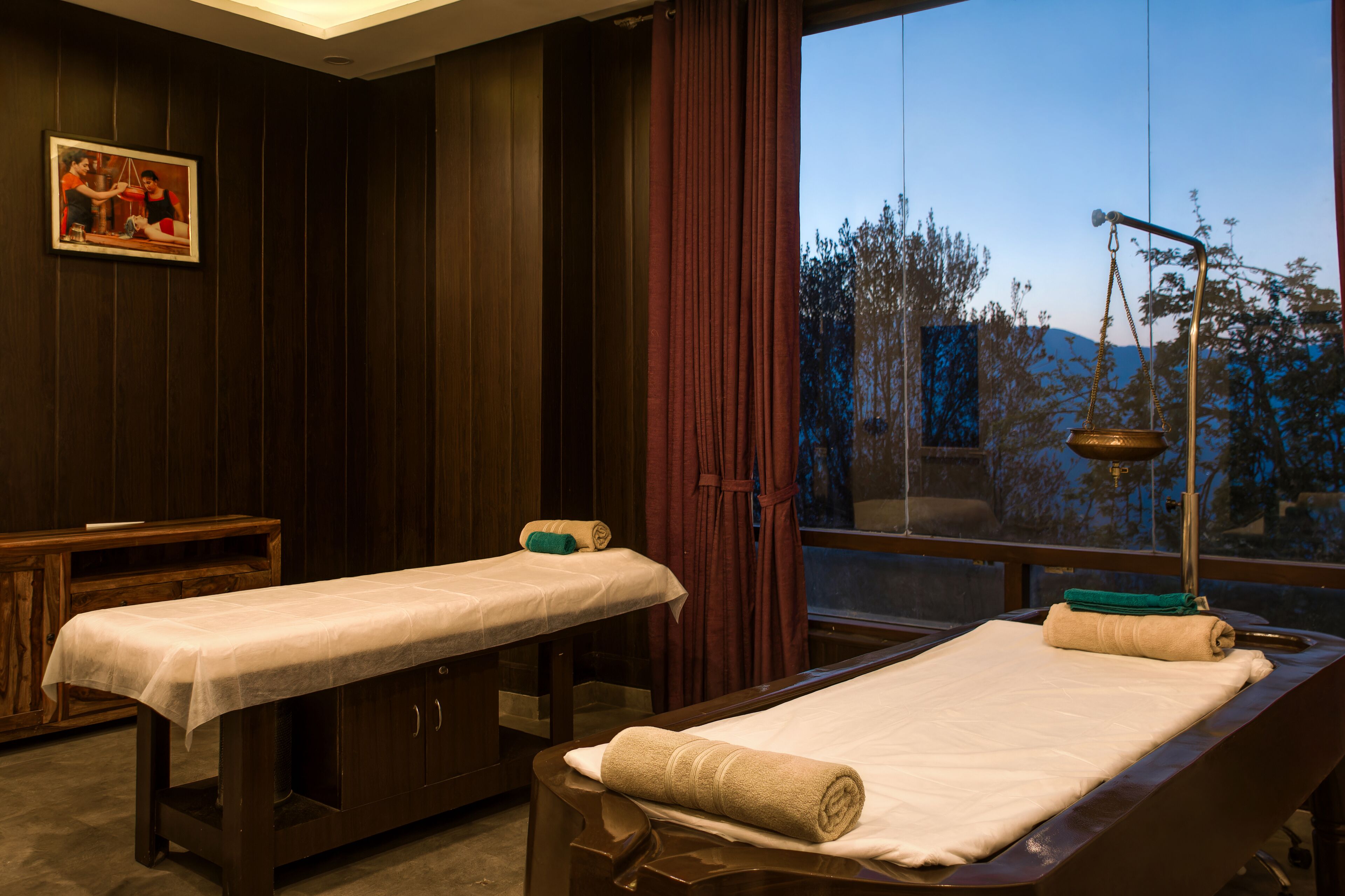 A room with two massage tables, a wooden cabinet, a framed picture, and a window with a view of trees and mountains.
