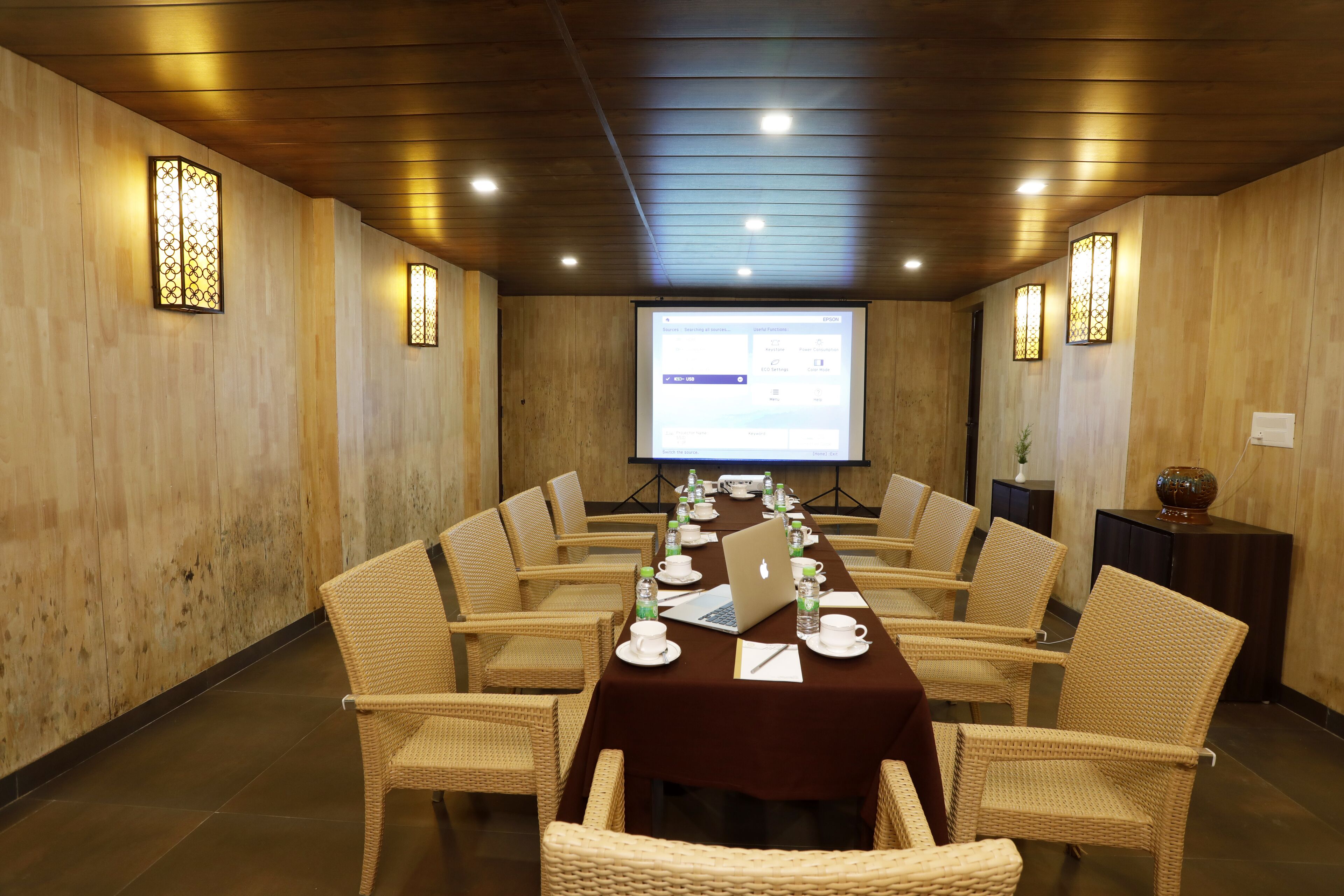 A conference room with a long table, wicker chairs, a laptop, and a projector screen.