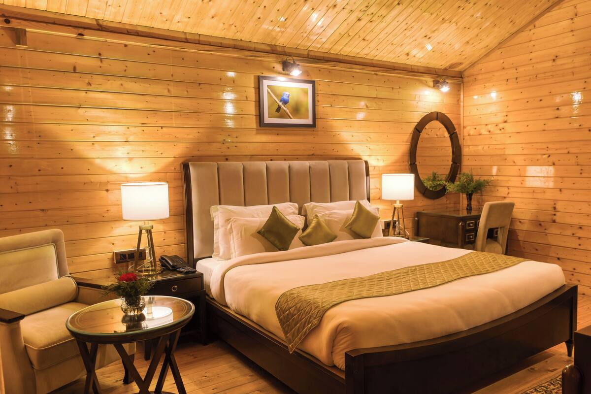 A wooden-paneled room with a bed, two bedside lamps, a chair, a small table with a vase, and a framed picture on the wall.