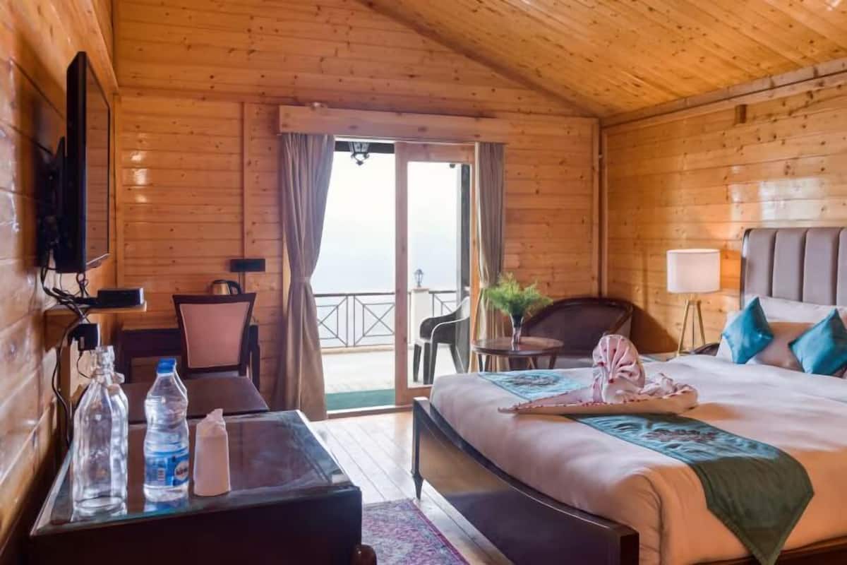 A wooden cabin room with a bed, a desk, a chair, a table with water bottles, and a TV.