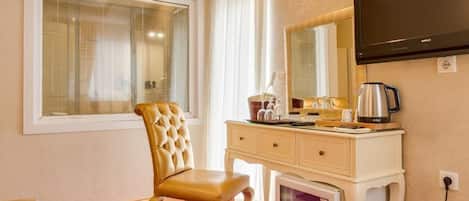 Habitación de hotel con una cama individual, una silla, tocador con espejo, televisión montada en la pared, cafetera y refrigerador.