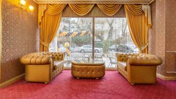 A luxurious room with a red carpet, two tufted sofas, a glass coffee table, and a view of the street through large windows with golden curtains.