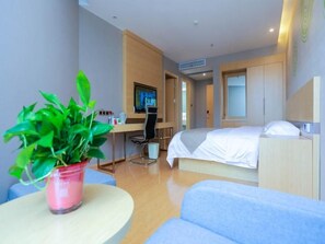 A modern hotel room with a bed, a desk, a chair, a TV, and a potted plant.