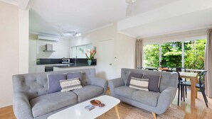 A modern living room with two grey sofas, a white coffee table, and a view of a kitchen and dining area with a large window.
