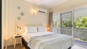 A bedroom with a bed, a nightstand, a wall-mounted air conditioner, starfish decorations, and a view of greenery through large windows.