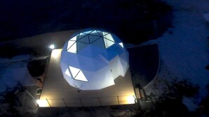 Un edificio esférico con diseño geométrico, iluminado desde el interior, ubicado sobre una superficie plana con cielo despejado al fondo.