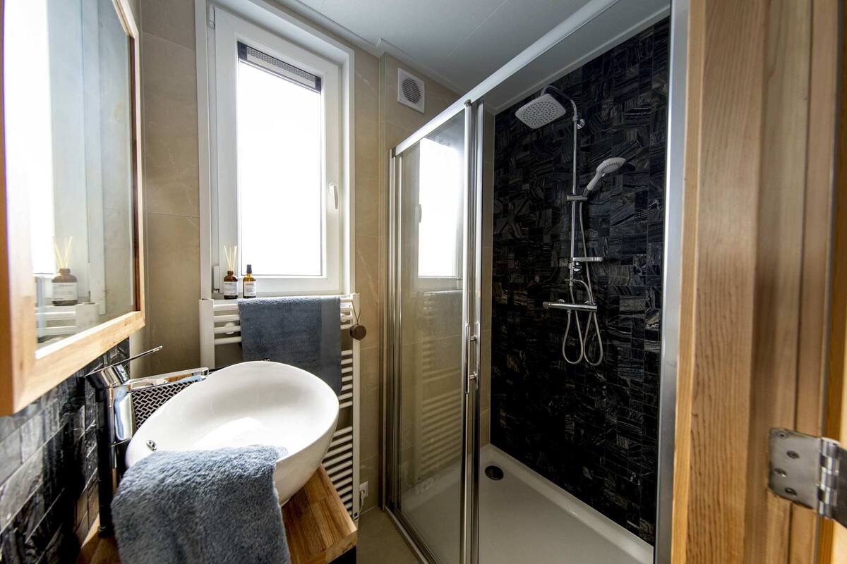Une salle de bain avec un lavabo blanc, une cabine de douche en verre et une fenêtre.