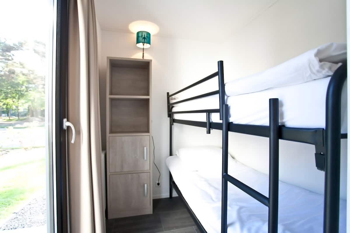 Un lit superposé avec une tête de lit, une commode en bois avec des tiroirs et un petit bureau avec une lampe.