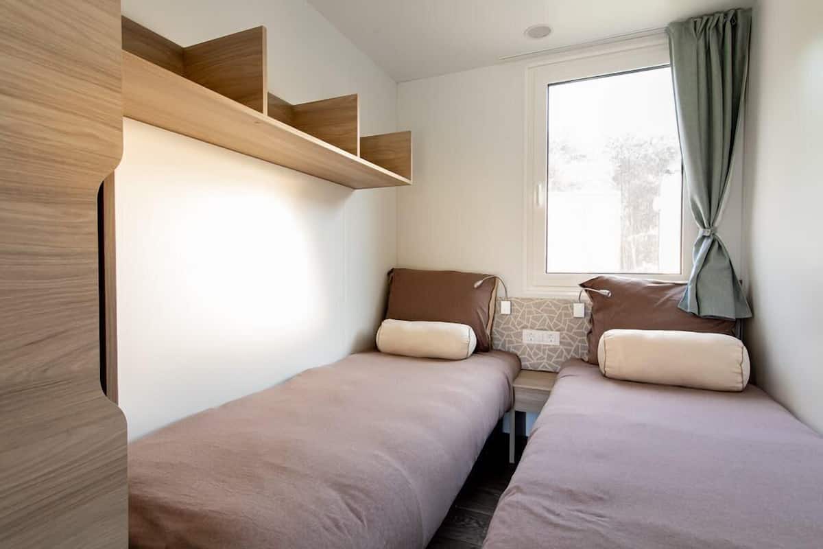 Une petite pièce bien éclairée, avec deux lits simples, une fenêtre avec des rideaux et des étagères en bois intégrées.