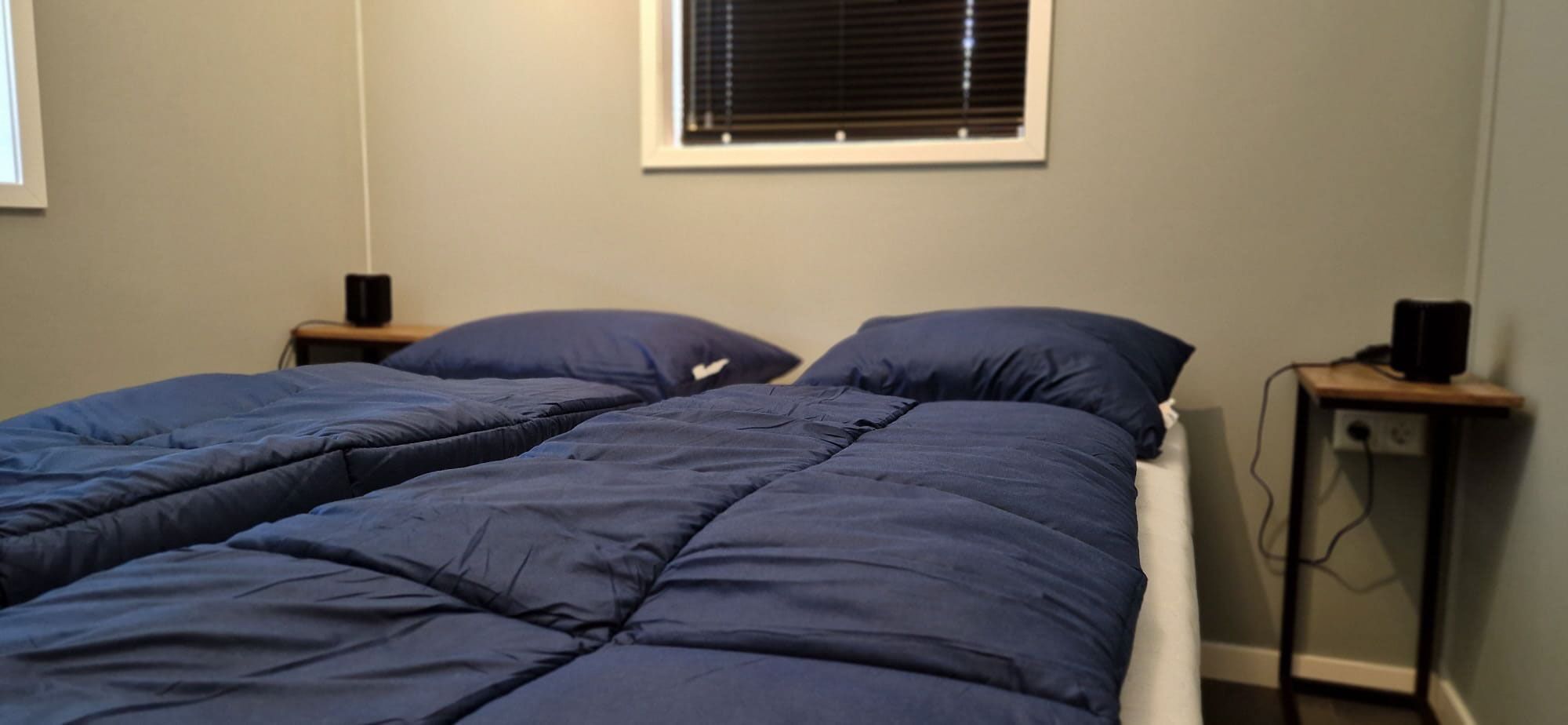 A twin bed with a blue comforter and pillows, a bedside table with a lamp, and a window with blinds.