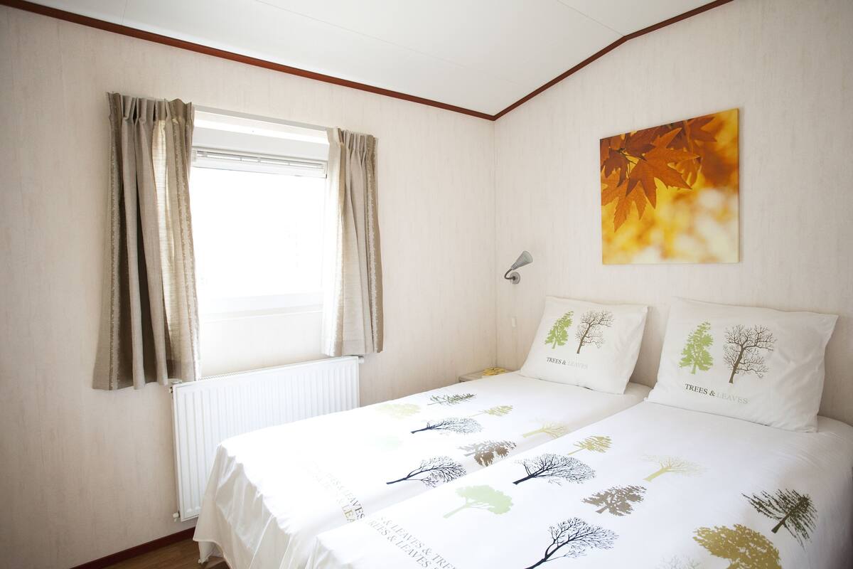 Une chambre à coucher avec un lit, deux oreillers ornés de motifs d’arbres, une fenêtre avec des rideaux et une lampe fixée au mur.