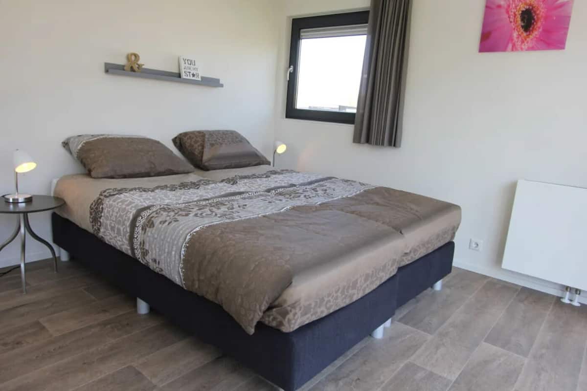 Une chambre à coucher comprenant un lit, des tables de chevet, une fenêtre avec des rideaux, un radiateur et une étagère fixée au mur, sur laquelle est posé un panneau.