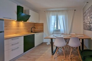 A modern kitchen with white cabinets, a wooden countertop, and a green backsplash. There is a dining area with a table and chairs, and a window with white curtains.