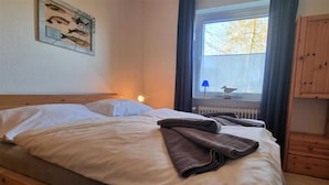 A double bed with white and brown linens, a framed fish artwork on the wall, and a window with a view of trees. - House Severin Whg Findling ground floor (Nieblum)