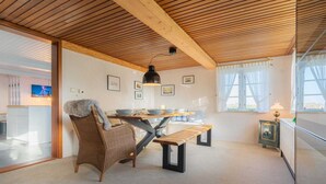 A dining area with a wooden ceiling, a wicker chair, a bench, a table with bowls, and a lamp. - Haus Husen - Haushälfte A Heini (Poppenbüll)