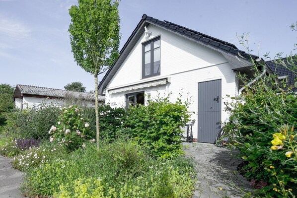 A white house with a grey door, surrounded by greenery and a bicycle. - Apartment Pesel - Vacation Rentals Northbeach Retreat (Nordstrand)