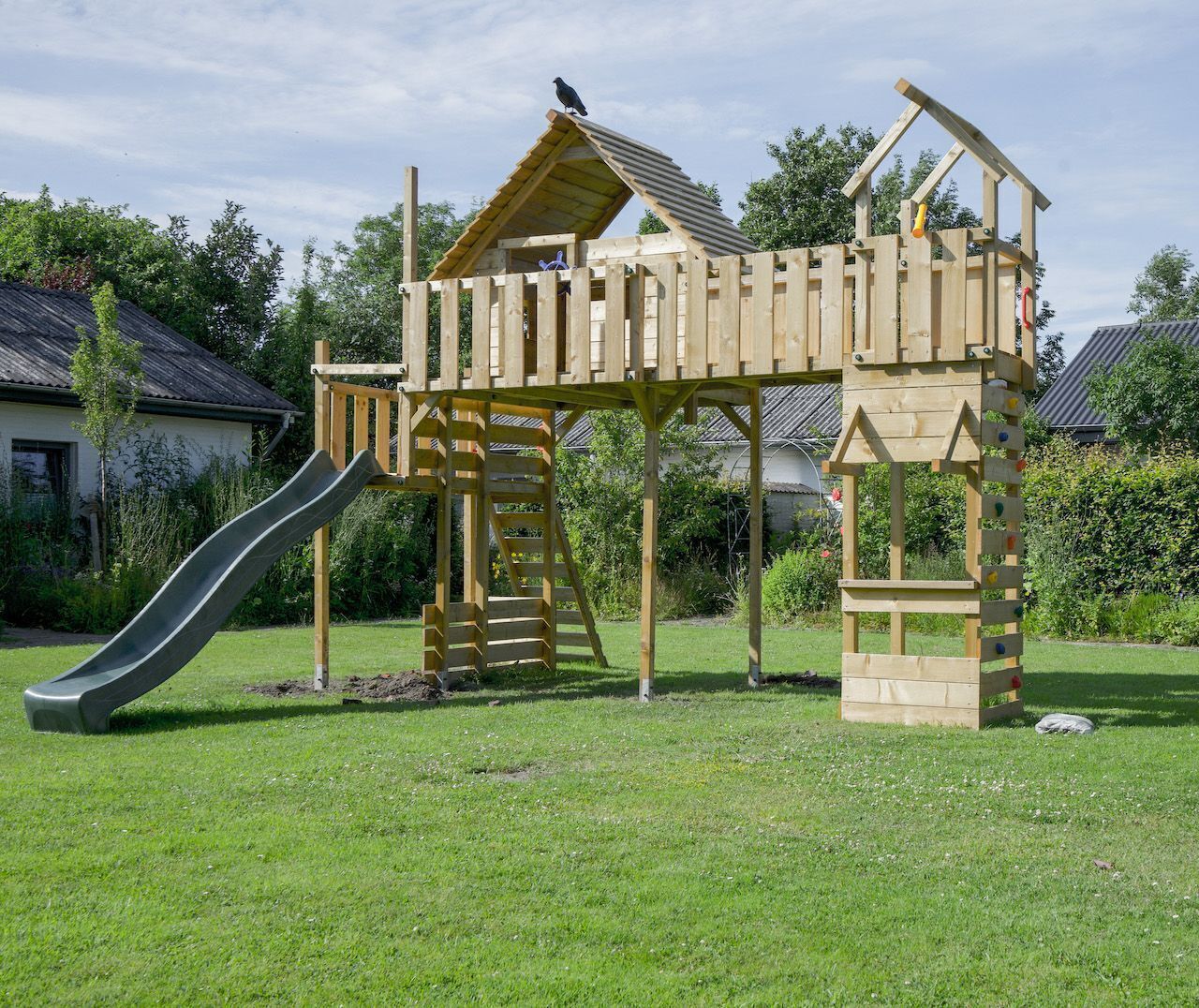 Une structure de jeu en bois, dotée d’un toboggan et d’un mur d’escalade, installée dans une cour gazonnée.