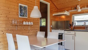Puinen mökki sisältä, jossa on ruokapöytä, tuoleja, keittiö liesineen ja tiskialtaineen sekä seinällä oleva kehystetty taideteos.