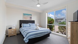 A bedroom with a bed, bedside tables, a ceiling fan, a window with a view, and a TV on a cabinet. - 4/27 South Street. Renovated three bedroom villa. (Tuncurry)