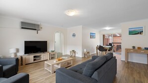 A modern living room with a flat-screen TV, grey sofas, a coffee table, and a dining area with a table and chairs. - 4/27 South Street. Renovated three bedroom villa. (Tuncurry)