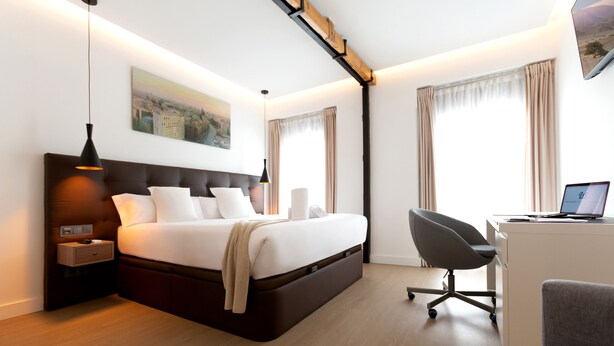 Una habitación de hotel moderna con una cama grande, mesitas de noche, un escritorio con una computadora portátil y un cuadro con una vista urbana en la pared.